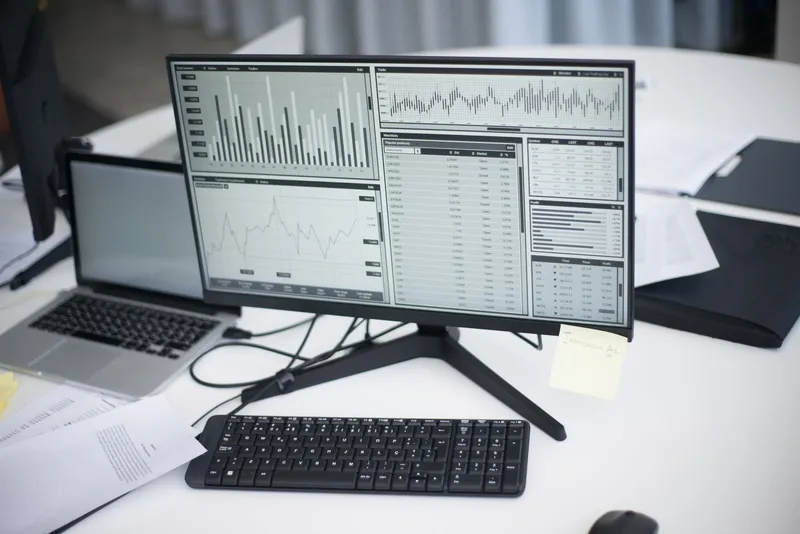 Cómo integrar datos de varias fuentes en un solo cuadro de mando: escritorio con un monitor y un portátil que muestran gráficas y tablas de datos, con papeles alrededor