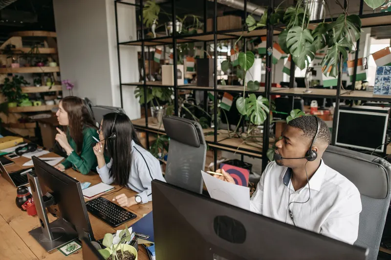 CRM con agentes de IA: qué es, cómo funciona y en qué se diferencia de un CRM tradicional: oficina con varias personas atendiendo llamadas con auriculares frente a sus pantallas, entre estanterías con plantas