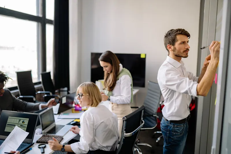 Software a medida o estándar: cómo decidir cuál necesita tu empresa: oficina en la que una persona escribe en una pared de cristal con notas adhesivas mientras el resto trabaja en la mesa